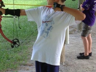 A young male camper stands with a bow and arrow drawn facing a target.