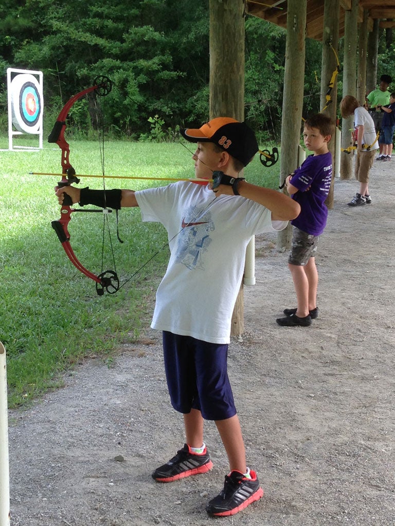 A young male camper stands with a bow and arrow drawn facing a target.