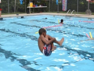 A young male camper does a cannonball into a pool. Other campers can be seen in the background floating on pool noodles.
