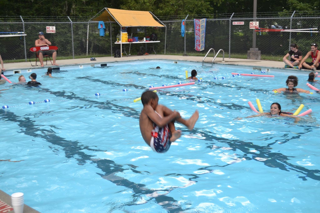 A young male camper does a cannonball into a pool. Other campers can be seen in the background floating on pool noodles.