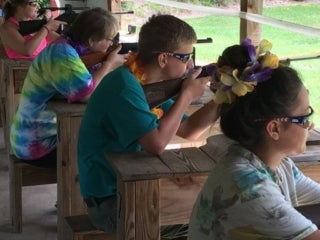 A group of campers sitting at a row of gun rests aiming long guns down a range.