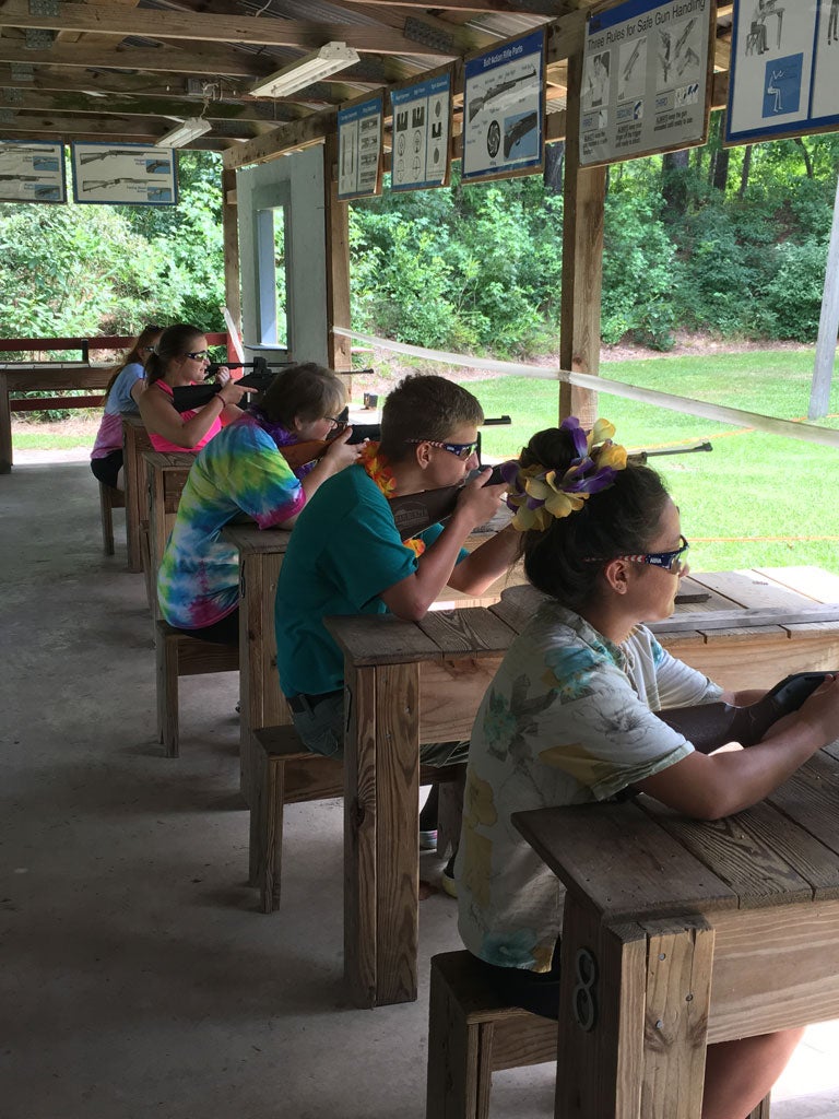 A group of campers sitting at a row of gun rests aiming long guns down a range.