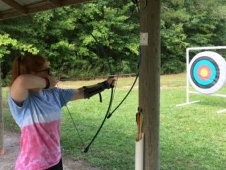 A female camper wearing a tie-dyed shirt holds a bow and arrow drawn facing a target.