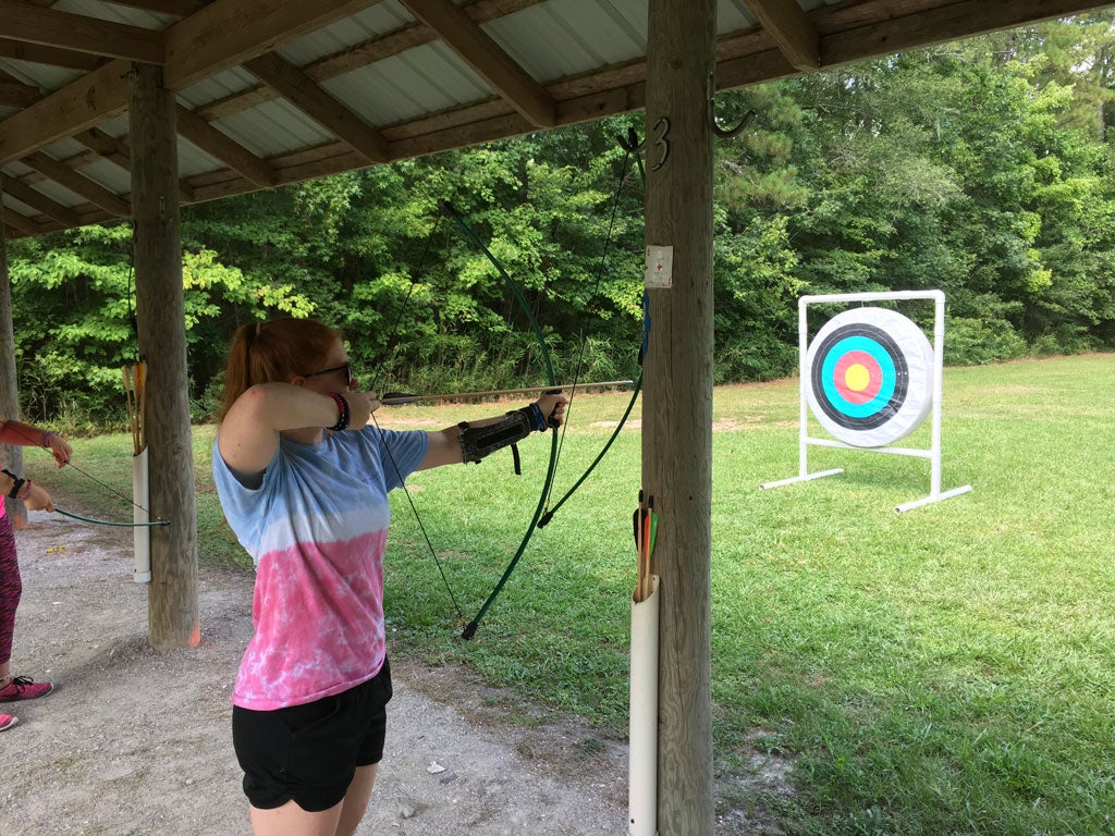 A female camper wearing a tie-dyed shirt holds a bow and arrow drawn facing a target.