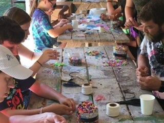 A group of campers sit around picnic tables working on a craft project with colorful beads.