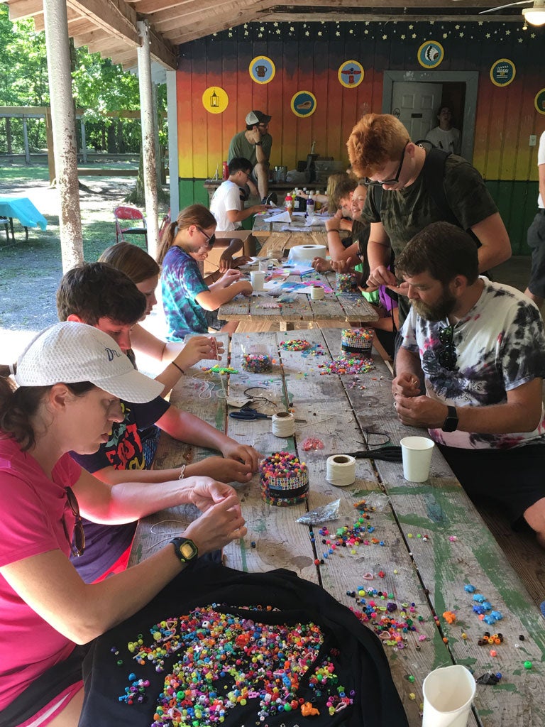 A group of campers sit around picnic tables working on a craft project with colorful beads.